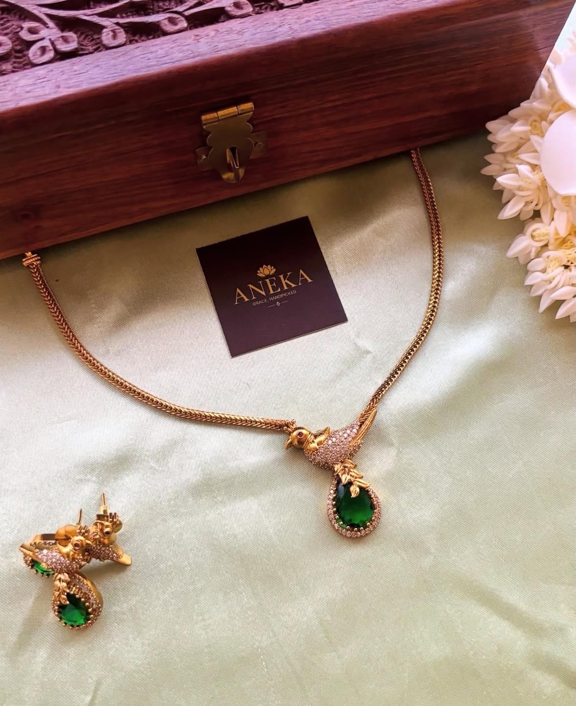 Elegant gold bird pendant necklace with green gemstone, paired with matching earrings, displayed on silk with wooden box.